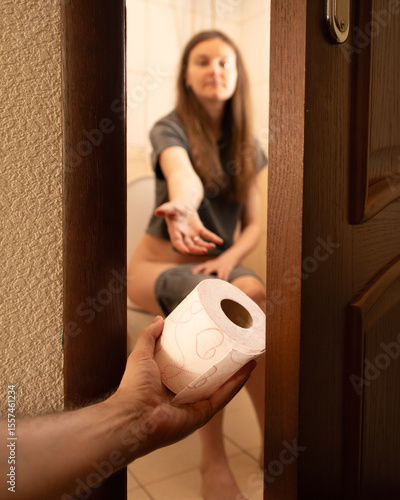 A person on the toilet reaches out for toilet paper while another hand offers assistance. This moment captures the everyday humor and necessity of shared spaces in a home