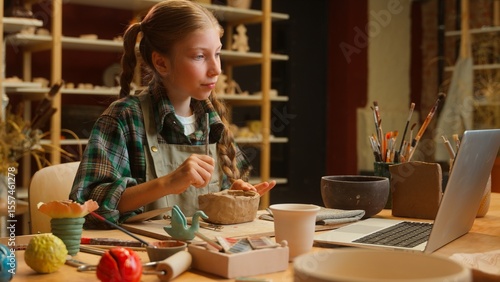 Laughing joyful Caucasian talented cute little girl teenager Caucasian schoolgirl child kid art school student sculpt clay modeling pottery ceramic artwork listen laptop video online tutorial indoors