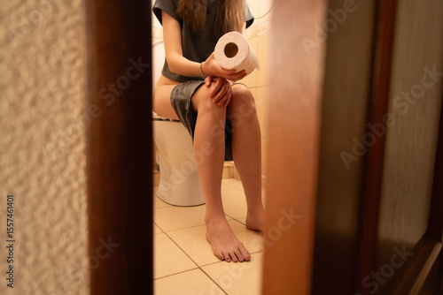 A person is sitting on a toilet with their pants down, holding a roll of toilet paper. The bathroom setting includes tiled floors and a simple design, emphasizing the daily routine