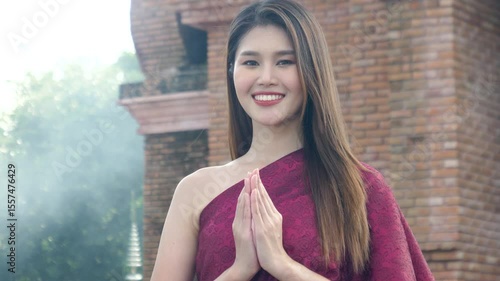 Smiling, beautiful young Asian woman wearing Thai traditional cloth greeting with Thai culture Sawasdee, welcome expression with temple in the background.