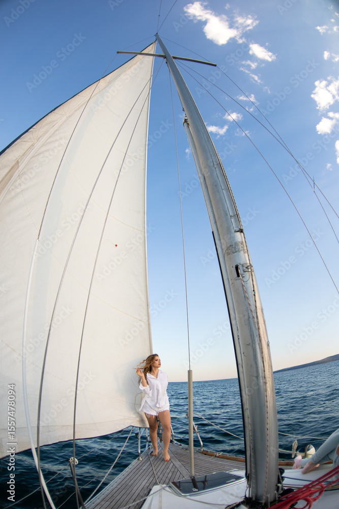 Fototapeta premium A woman in a white shirt relaxes aboard a sailing yacht, its tall white sails billowing above the deck as calm blue ocean stretches around her
