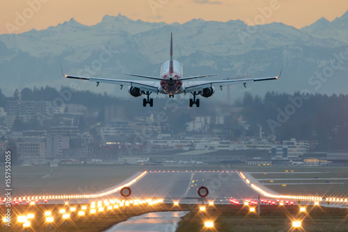 Commercial airliner landing at dusk on illuminated runway with city and mountains in background, aviation photo of jet airplane on approach with landing gear down at twilight