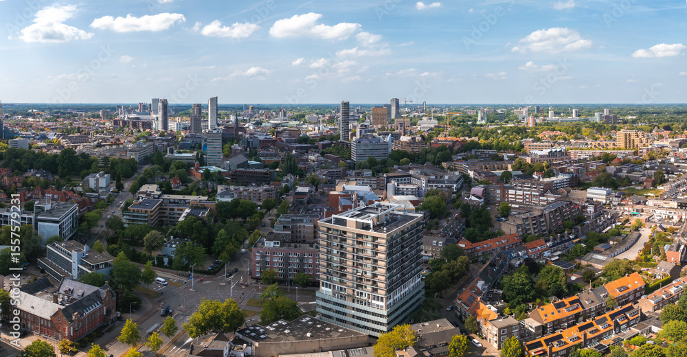 Obraz premium Aerial view of Eindhoven, Netherlands, featuring high rise buildings, red roofed houses, tree lined streets, green spaces, and a partly cloudy sky.