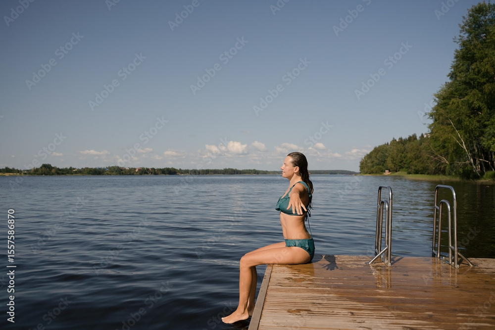 Fit woman in a swimsuit splashing water with her feet while sitting on a pier. Relaxed moment of summer and wellness. Fit woman in a swimsuit splashing water with her feet while sitting on a pier. Relaxed moment of summer and wellness.