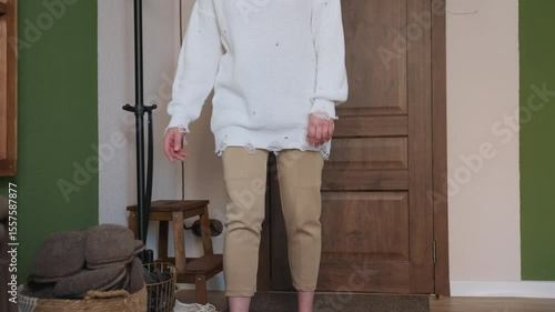 Patient stands in cozy room after removing canvas shoes, preparing to wear crocs, wearing beige pants and oversized white sweater, in front of wooden door with green walls, and indoor furniture