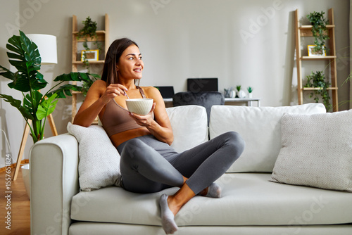 Young athletic woman eating healthy breakfast at home sitting on sofa