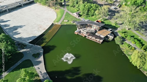 Angled drone video in arc motion around Robinson Restaurant and Városligeti-tó lake in Budapest, with a partial view of Zielinski Bridge and surrounding greenery in morning light. 