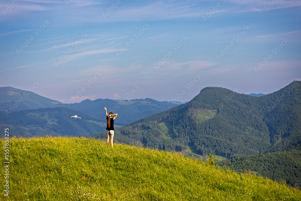 Fototapeta premium Drone Filming Girl on Scenic Mountain Hill. Young woman standing on a grassy hilltop posing for a drone camera, with vast green mountains and clear sky in the background.