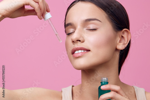 Εκτύπωση καμβά Young woman applying facial serum with a dropper on a pink background, showcasin
