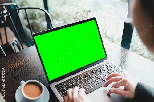 Over the shoulder view of a person using a laptop with a blank green screen in a cafe. A chroma key mockup for video or website demonstrations.