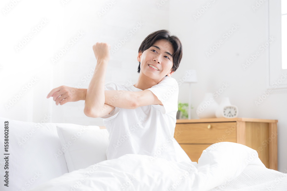 朝日を浴びるベッドで寝起きの若い男性 腕を伸ばしストレッチをする 朝日を浴びるベッドで寝起きの若い男性 腕を伸ばしストレッチをする