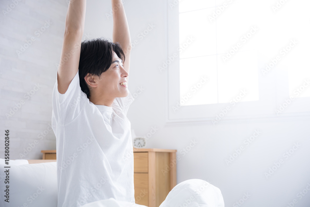 朝日を浴びるベッドで寝起きの若い男性 腕を伸ばしストレッチをする 朝日を浴びるベッドで寝起きの若い男性 腕を伸ばしストレッチをする