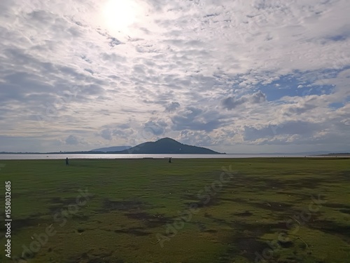 Scenic wide-angle shot of a lush green field by a calm lake, with a mountain in the distance under a dramatic cloudy sky. A peaceful nature scene capturing open space and atmospheric light.