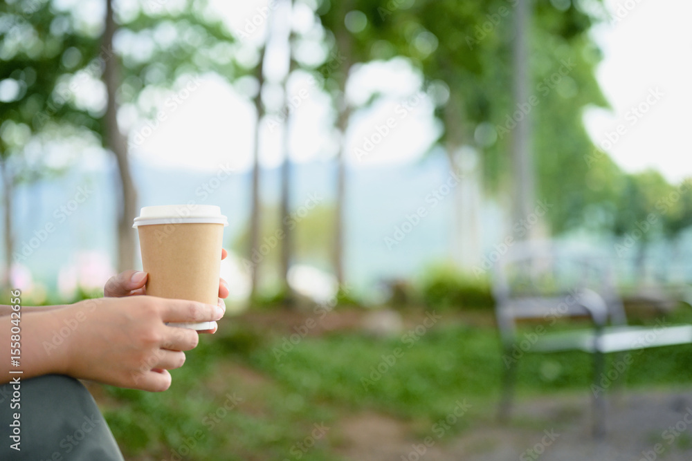 Close up of hands holding a paper coffee cup outdoors with a peaceful, green park background Close up of hands holding a paper coffee cup outdoors with a peaceful, green park background