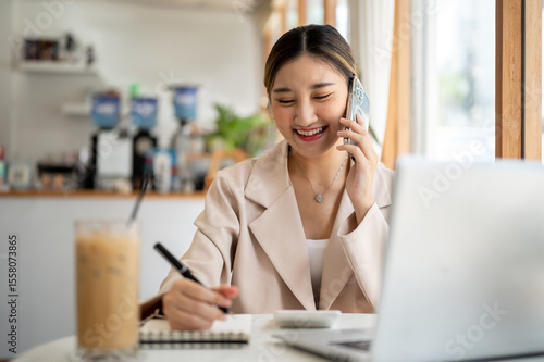 Asian woman talking on phone with pen on notebook as sitting with laptop and coffee at cafe's table.