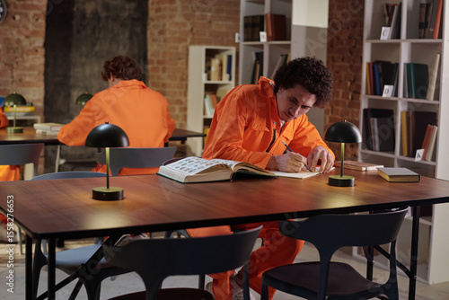 Young adult man wearing prison uniform sitting at table in prison library studying open book and writing notes while another young adult man reading in background