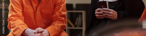Caucasian man in orange prison uniform sitting next to Caucasian woman dressed as nun holding book with cross, both seated in prison library setting