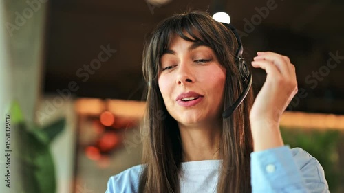 Cheerful professional businesswoman wearing a light blue shirt over a white t-shirt, actively engaged in online communication from a modern office, smiling happily.