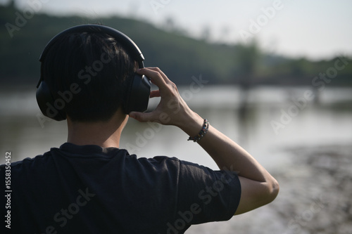 Fotografi A person wearing large headphones listens to audio while standing outdoors near a calm body of water with a blurred forest background