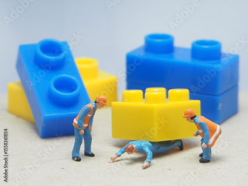 Mini toy of action figure and block at table with blurred background. Diorama photography concept design. Minifigure of worker character trapped under the block. Accident at site plant. Safety work.