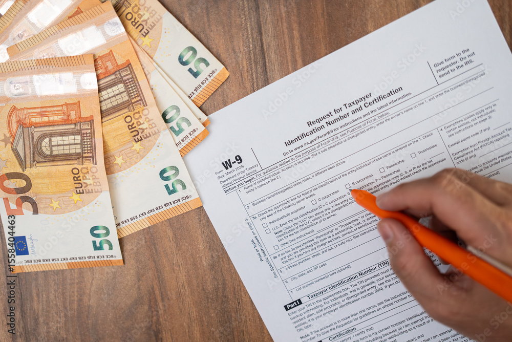 Fototapeta premium Independent contractor sits at wooden table with euro bills with pen in hand filling W-9 form to confirm TIN for reporting income from various business clients