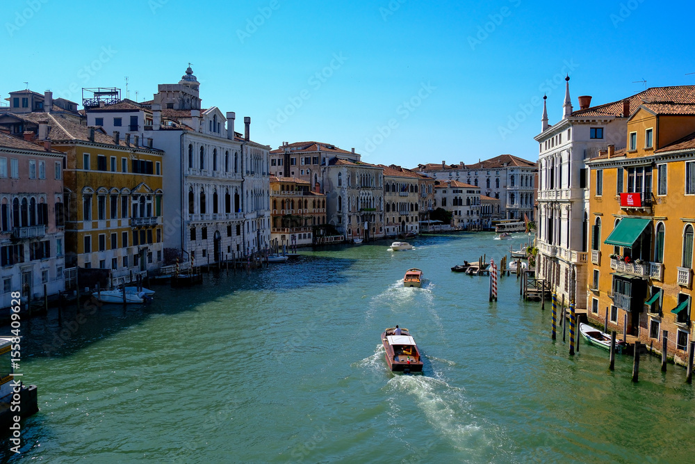 Fototapeta premium Lively Venetian canal with arched pedestrian bridge, colorful facades, waterfront cafés and boats under a bright blue sky – authentic travel scene showcasing everyday charm of Venice’s city life