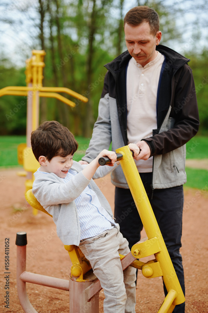 Fototapeta premium father and son enjoying outdoor fitness equipment in park. man guiding child on yellow exercise machine. family time promotes healthy lifestyle and bonding moments in nature