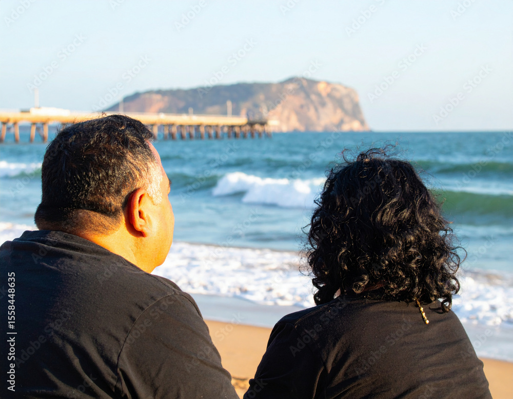 Fototapeta premium A couple sits on a sandy beach, gazing at the ocean waves and distant cliffs, enjoying a peaceful moment together.