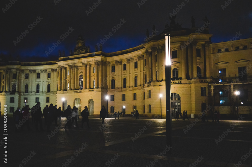 Fototapeta premium Night view of Bebelplatz square in Berlin, Germany, featuring illuminated historic buildings, silhouettes of people walking, and atmospheric city lights.
