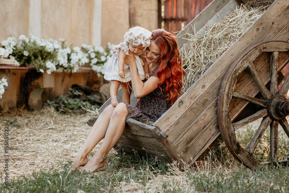 Obraz premium Happy young mother with baby in her arms sits in cart at sunset in village in summer. Girl with red hair. Motherhood. Mother's day. Mom and daughter