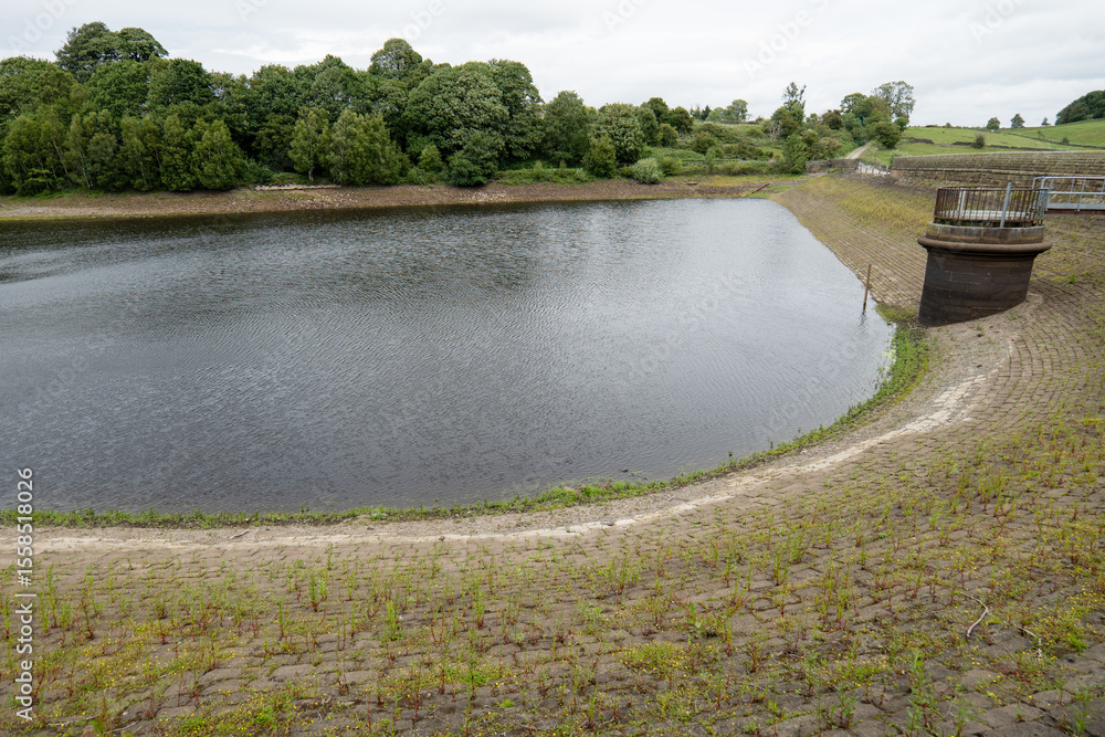 A depleted reservoir in the Yorkshire countryside shows low water levels exposing stone embankments in the rural English landscape. A depleted reservoir in the Yorkshire countryside shows low water levels exposing stone embankments in the rural English landscape.