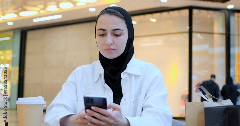 Fototapeta premium Young entrepreneur wearing hijab reviews notes and uses tablet in bustling shopping center food court, showcasing modern work practices and cultural diversity. Muslim diligent student, flexible