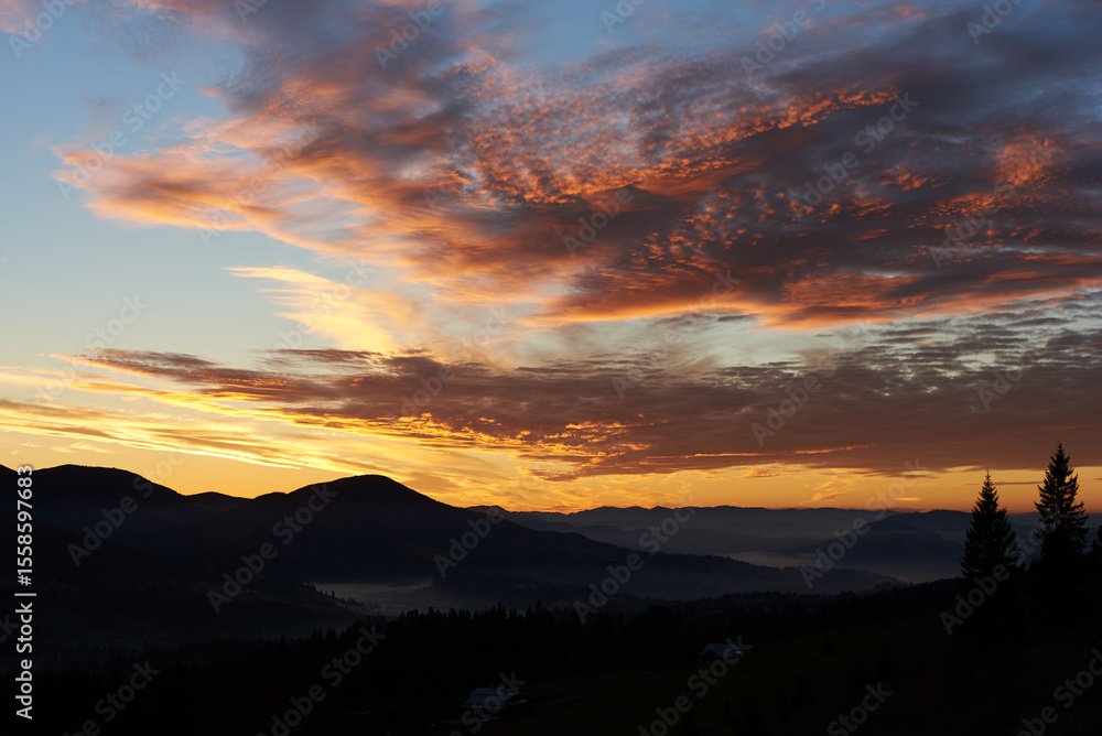 Fototapeta premium Breathtaking sunset paints sky with vibrant hues of pink, orange, purple, blending into blue morning sky. Silhouetted mountains, pine trees create serene backdrop. Tranquil and picturesque landscape.