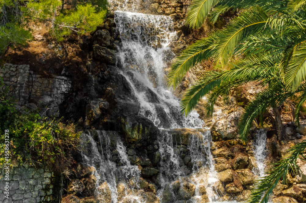Naklejka premium Scenic Artificial Waterfall Surrounded by Palm Trees and Rocks in Loutraki, Corinth, Greece, on a Bright Summer Day.