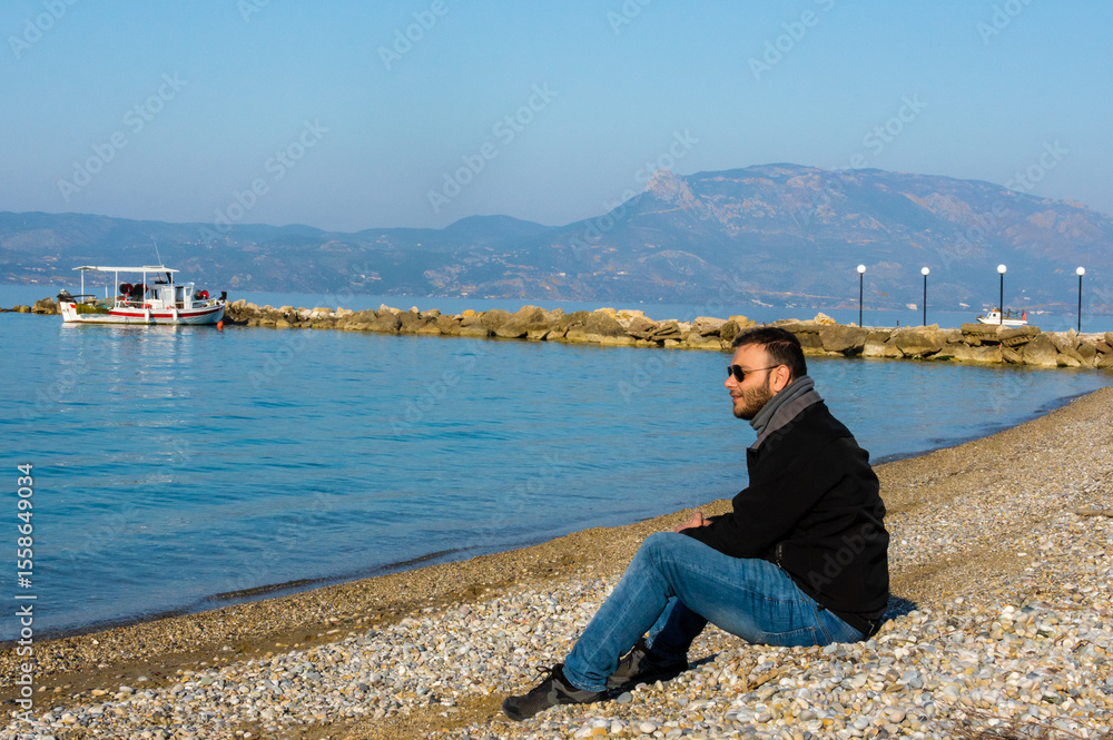 Fototapeta premium Relaxed Young Man Sitting on a Pebble Beach in Kato Assos, Corinth, Greece, with a Peaceful Sea View and Natural Light in a Fishing Village Setting.