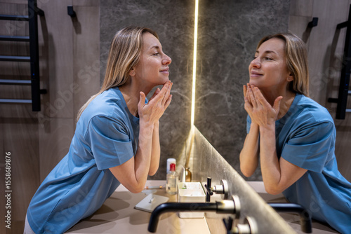 Fotografija Confident middle-aged woman looks to reflection in bathroom mirror touching face with smile