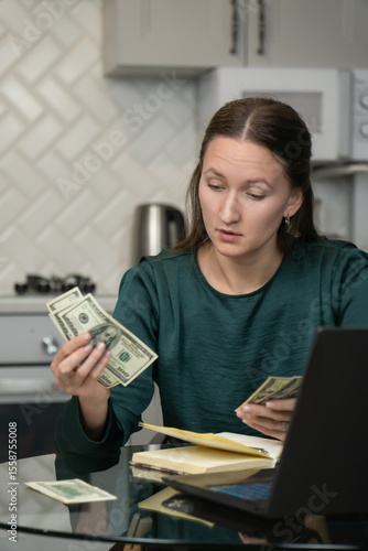 Young woman managing home finances, carefully counting us dollar banknotes while sitting at kitchen table with laptop and notebook, demonstrating financial responsibility and household budgeting