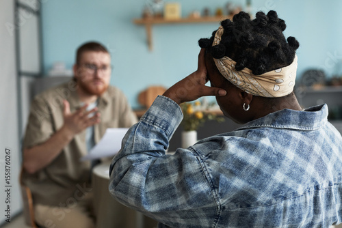Slika na platnu Black woman with headscarf sitting with back to camera, holding head while Cauca