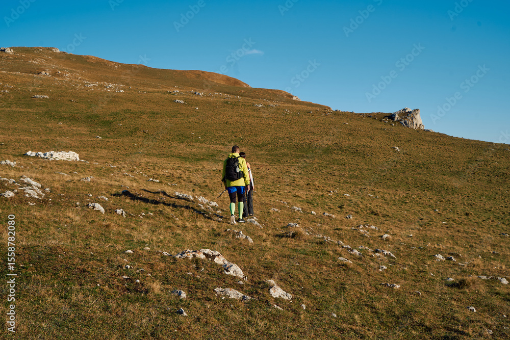 Fototapeta premium Couple hikers trek up a rocky mountain trail, enjoying a scenic view above the clouds, with majestic peaks in the background.