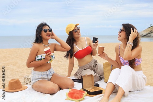 Three happy young Asian women enjoying pizza and drinks during a beach picnic on a sunny summer day. Group of friends relaxing on vacation, laughing and having fun outdoors near the sea.