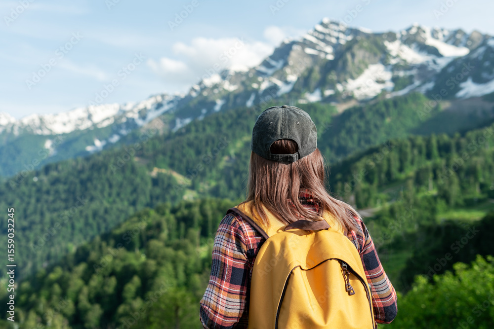Naklejka premium Young woman with a yellow backpack stands in front of majestic mountains, surrounded by lush greenery, enjoying the breathtaking view of nature's beauty and tranquility