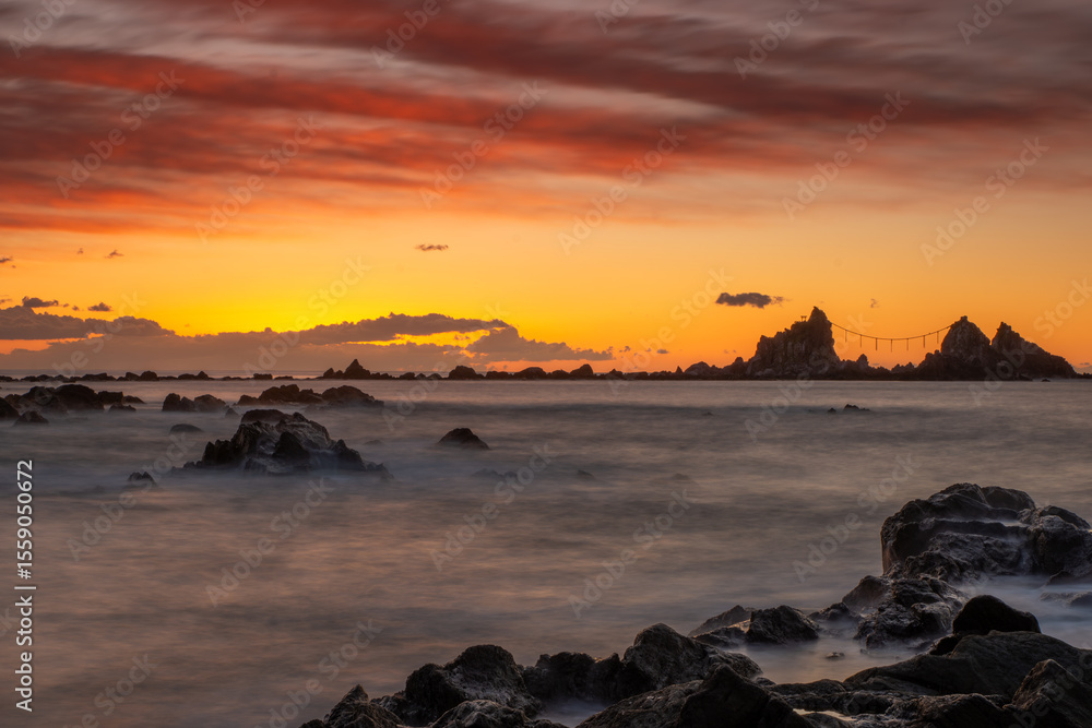 Fototapeta premium 神奈川県真鶴町の真鶴岬で撮影した三ツ石と朝焼けの海岸風景。オレンジ色に染まる空と静かな海、岩礁が印象的な冬の絶景。パワースポットや観光名所としても知られ、旅や癒しのシーン、和のイメージ素材に最適です。