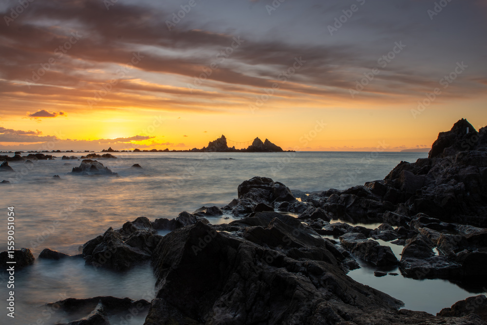 Fototapeta premium 神奈川県真鶴町の名勝・真鶴岬三ツ石に昇る朝日。オレンジ色の朝焼けと波打つ岩礁海岸が幻想的な絶景を創出。冬の澄んだ空気と静寂に包まれた神秘的な景色は、旅行・観光・自然風景・パワースポットのイメージに最適です。