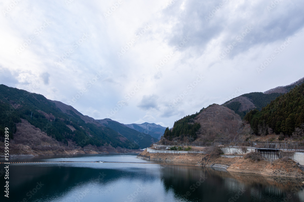 Obraz premium 埼玉県秩父市の滝沢ダムによってできたダム湖「奥秩父もみじ湖」の冬の風景。曇り空の下、静まり返った湖面に冬の山々が映り込む、荘厳で美しい景色。日本の山間部の穏やかな自然。縦横両方の構図あり。