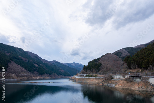 埼玉県秩父市の滝沢ダムによってできたダム湖「奥秩父もみじ湖」の冬の風景。曇り空の下、静まり返った湖面に冬の山々が映り込む、荘厳で美しい景色。日本の山間部の穏やかな自然。縦横両方の構図あり。

