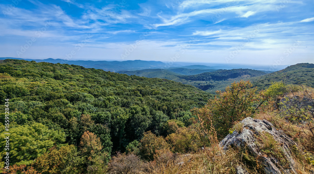 Naklejka premium Looking southwest from Őr-kő, this scenic landscape in the Bükk Mountains captures early signs of autumn across the forested terrain. Rolling hills meet open sky, framed by ancient trees. 