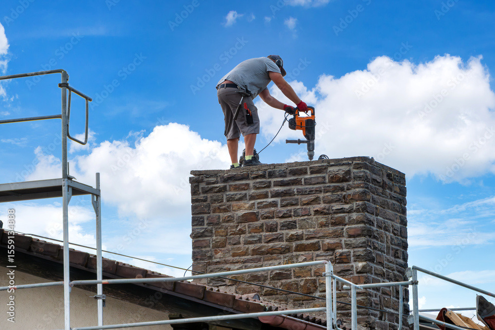 Naklejka premium Worker with jackhammer starts demolishing a brick chimney on the roof of a residential building