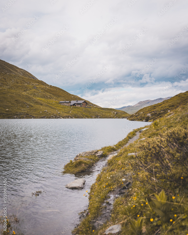 Fototapeta premium Mountain lake with hiking trail and alpine refuge under a moody sky in summer landscape