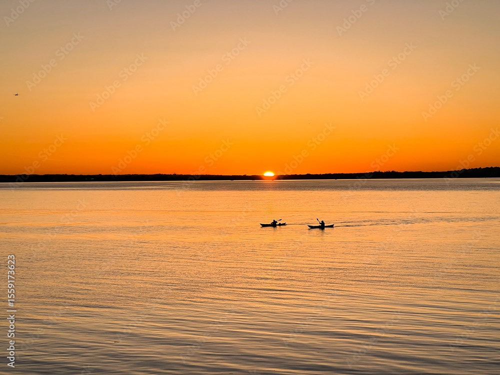 Naklejka premium Silhouettes of kayakers paddling across peaceful sea as the sun sets behind the distant horizon