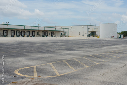 Empty Industrial Lot with Warehouse and Storage Facility in the Background. Empty paved area in front of a warehouse building and storage tank under a clear sky.
